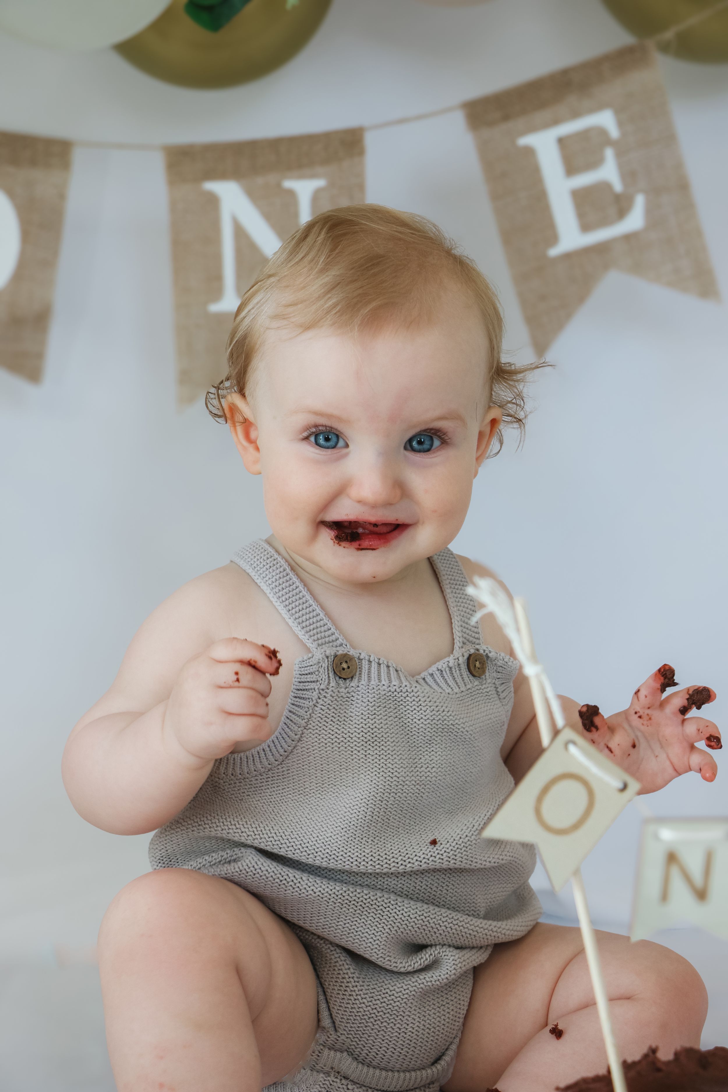 Smiling baby wearing gray knit romper sitting against banner backdrop with purple jelly or cake smeared on face.