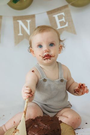 Baby in gray romper with chocolate cake smeared on face during first birthday celebration under banner decorations.