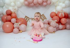 Baby in pink dress sits with smash cake surrounded by coral and white balloon arch for first birthday celebration.