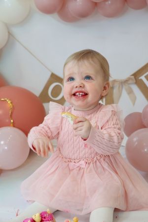 Baby wearing pink knit dress smiles while sitting against backdrop of pink and white balloons at birthday celebration.