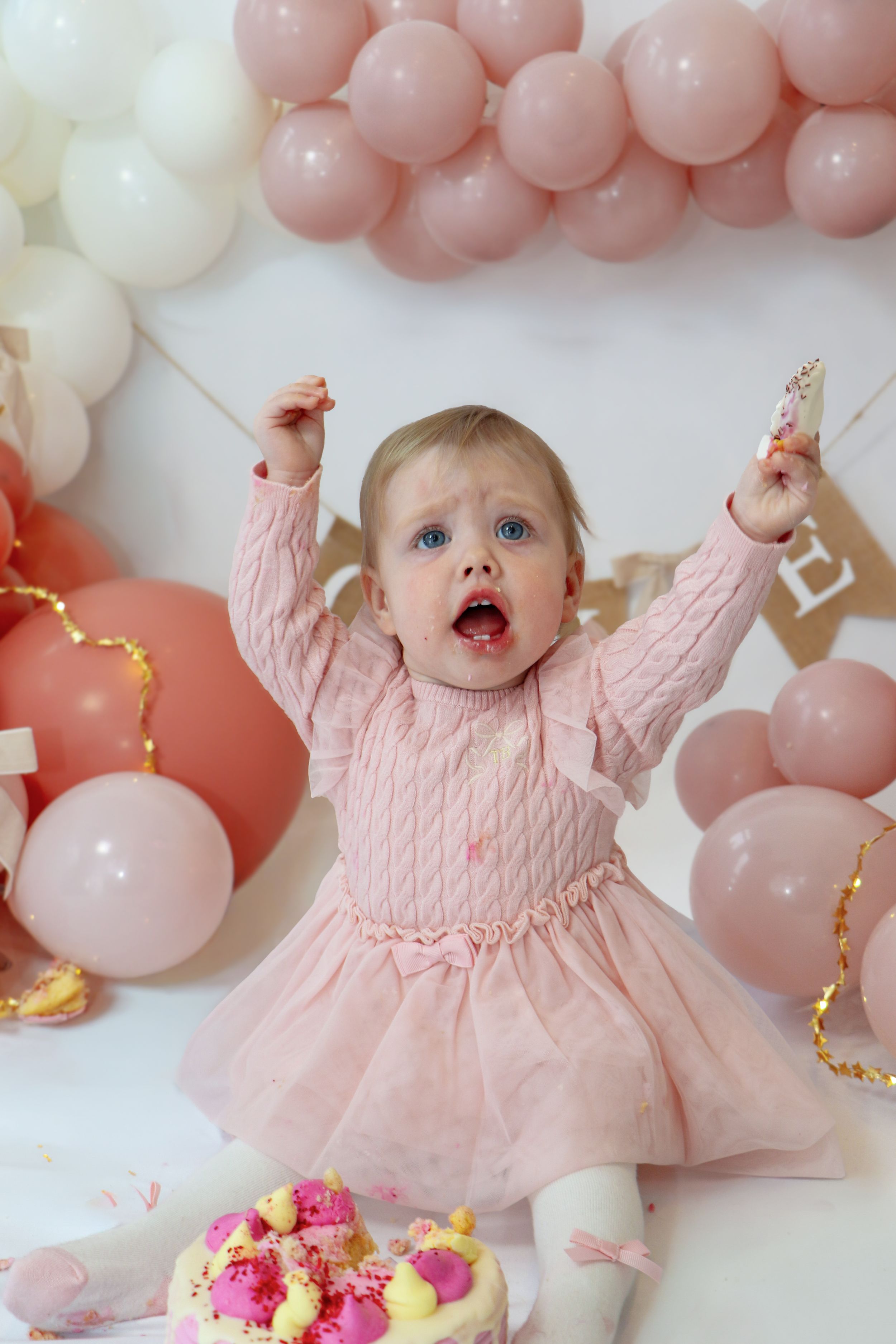 Baby in pink knit dress celebrates with pink and white balloon decorations and floral accents.