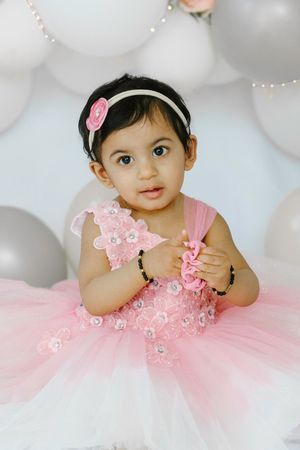 A small child in a pink tulle dress with floral details poses against a backdrop of white balloons for a portrait session.