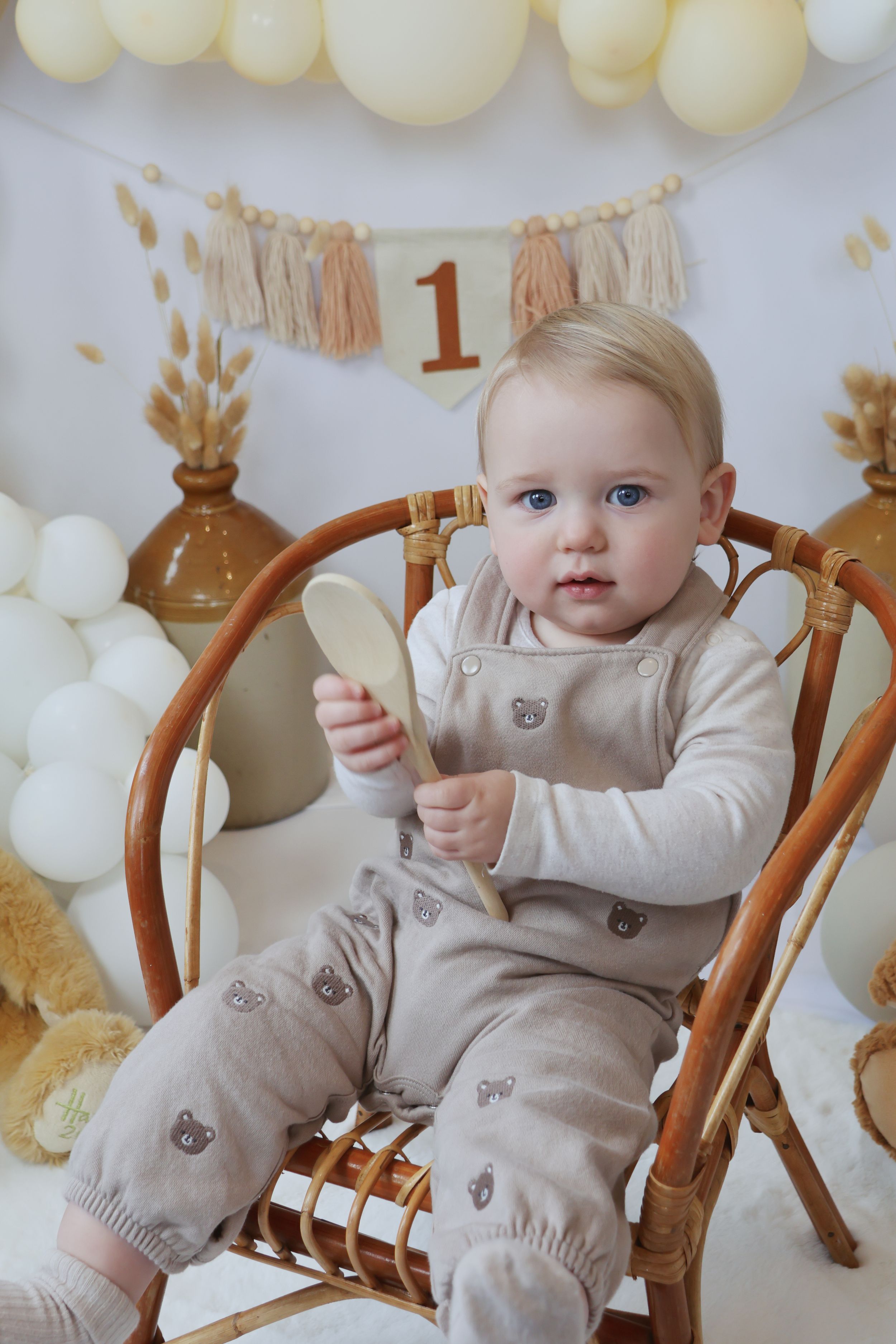 A baby in a beige outfit sits in a wooden chair surrounded by white and gold party decorations for a first birthday.