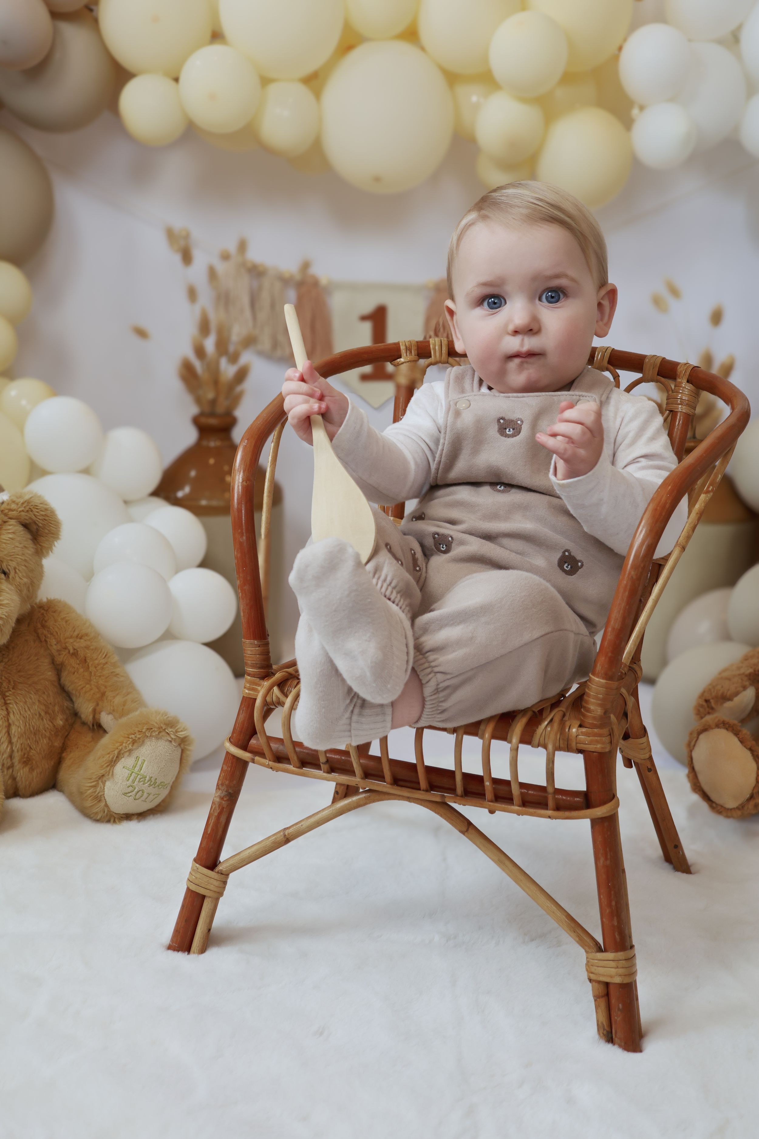 Baby sits in wooden rocking chair with white balloon backdrop and teddy bear decorations for first birthday photo shoot.