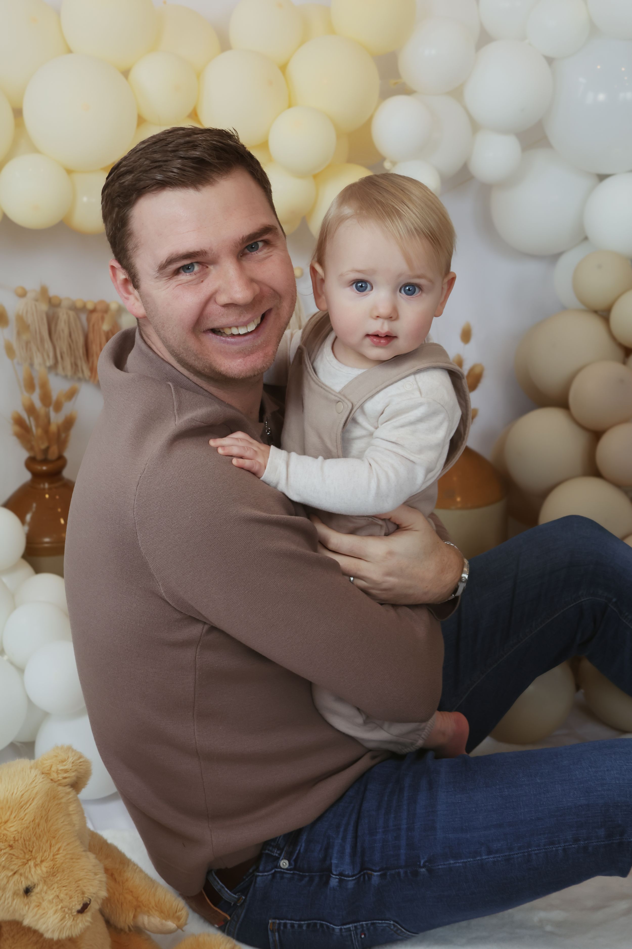 A parent and toddler pose together against a white balloon backdrop with teddy bears for a festive portrait session.