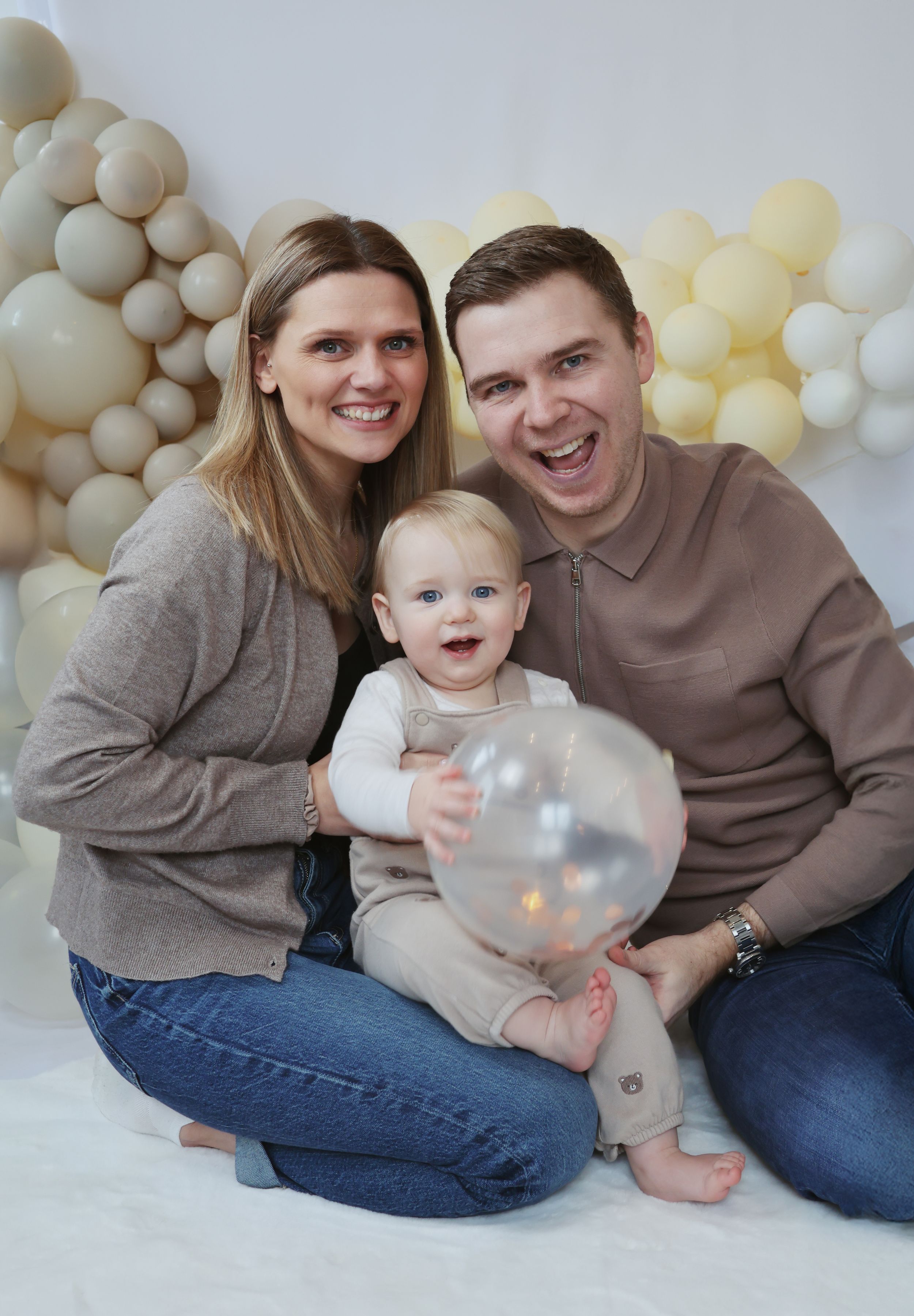 Family in matching gray sweaters poses together with white balloons and soft lighting for a cozy studio portrait session.