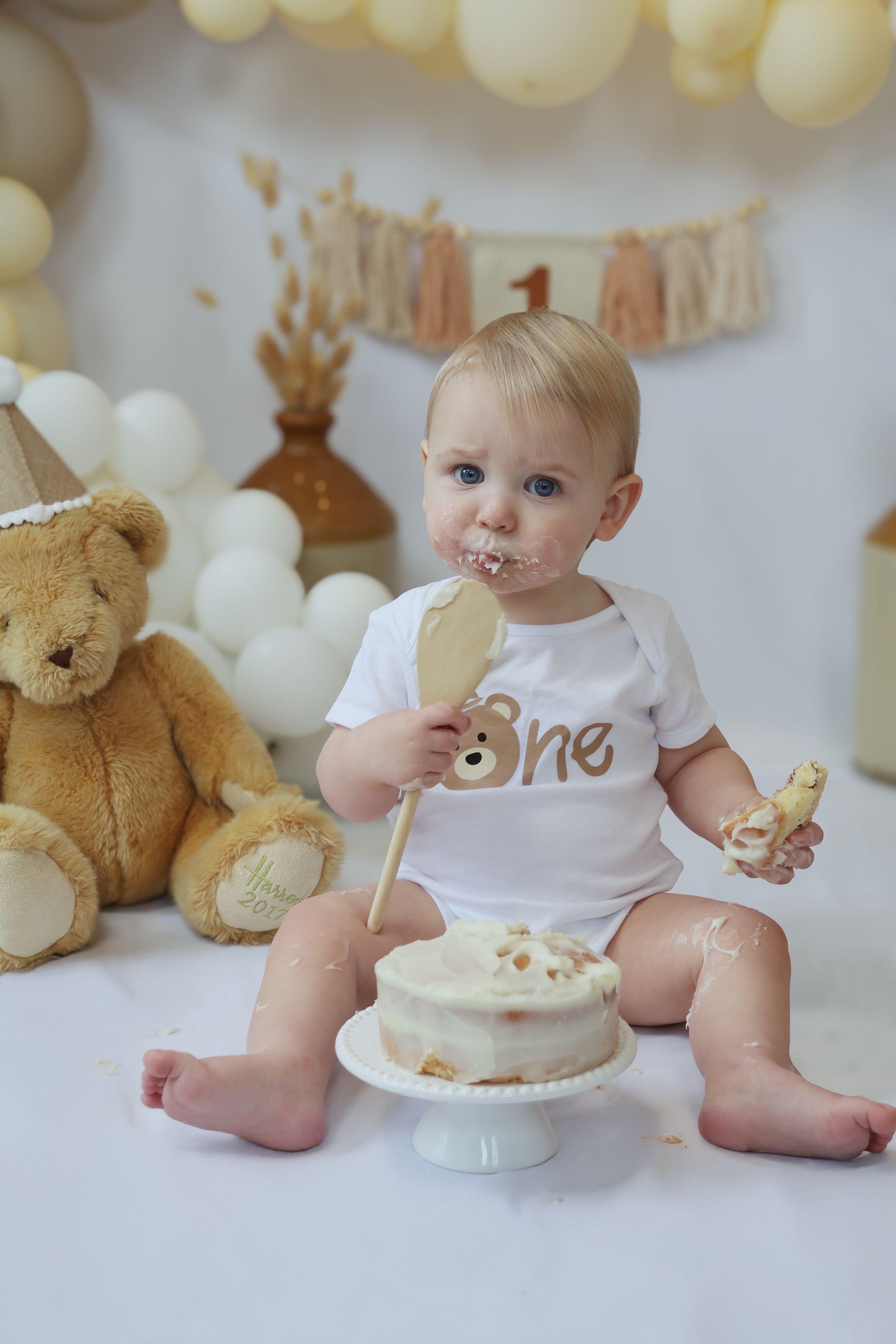 First birthday cake smash photo setup with teddy bear, white balloons and banner decorations for milestone celebration.