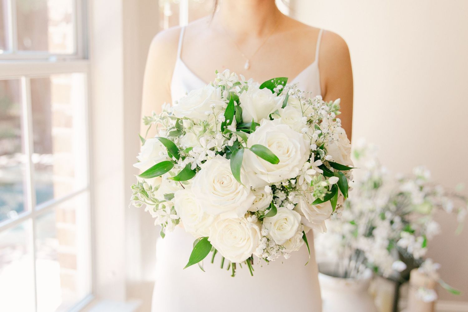 A bride holds a delicate white flower bouquet with a pink ribbon against a light, bright background.