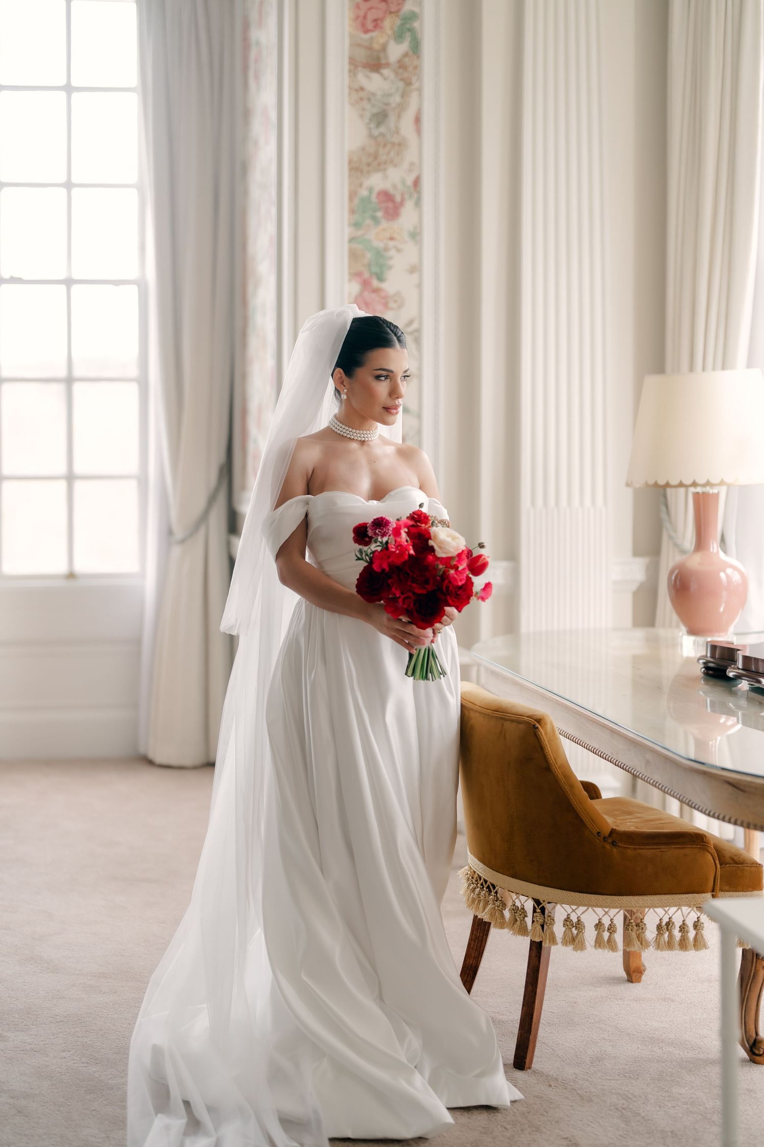 Bridal portrait session in a bright sunlit room with red bouquet and vintage velvet chair.