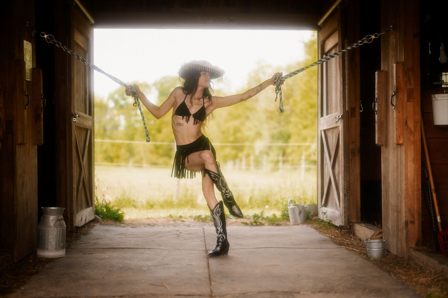 A dancer in western-style costume and cowboy boots performs an elegant pose inside a rustic barn entrance.