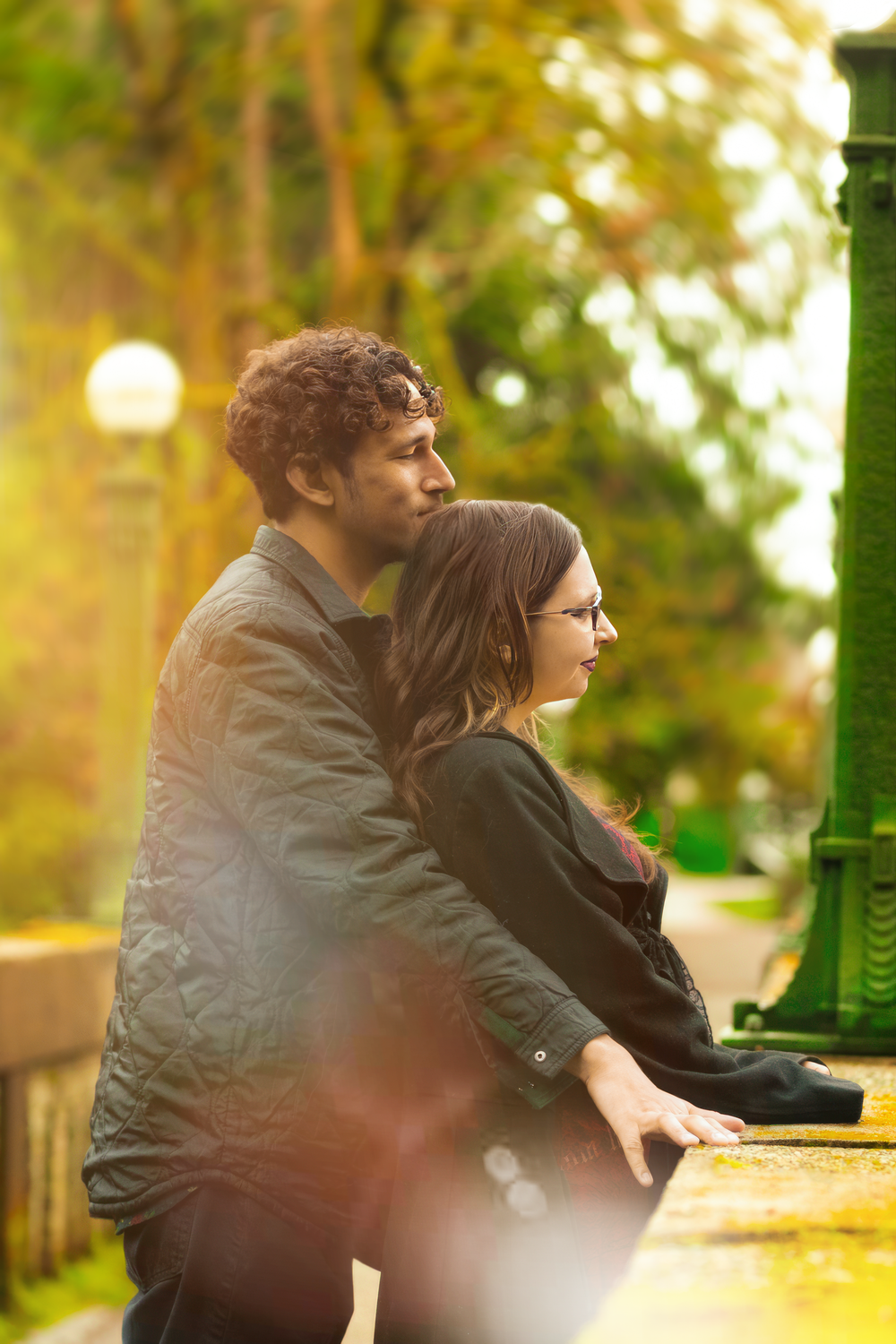 Couple standing side by side at railing during golden sunset in autumn park with soft warm lighting.