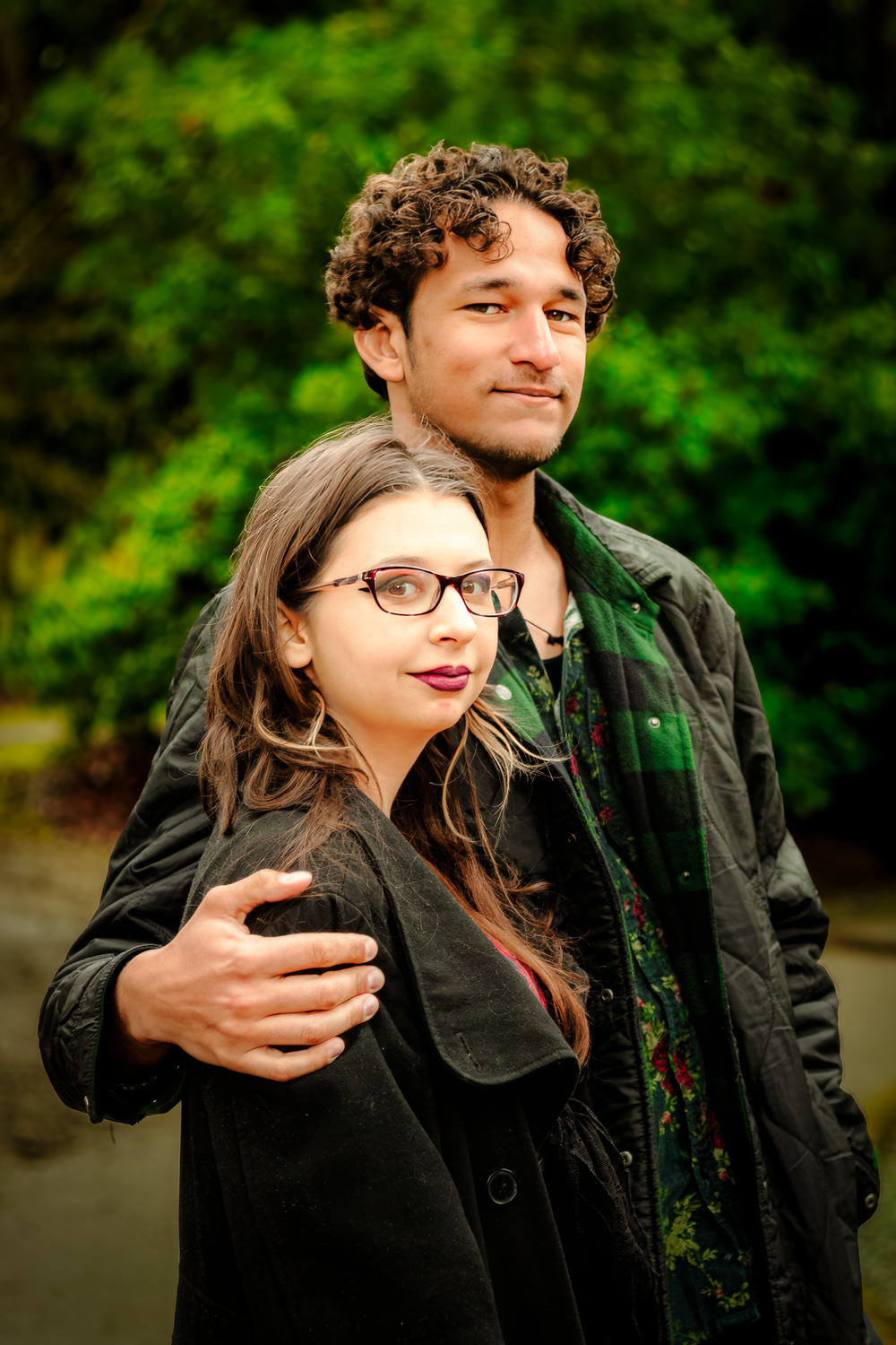 A couple in dark jackets embraces outdoors against a green natural background on an overcast day.