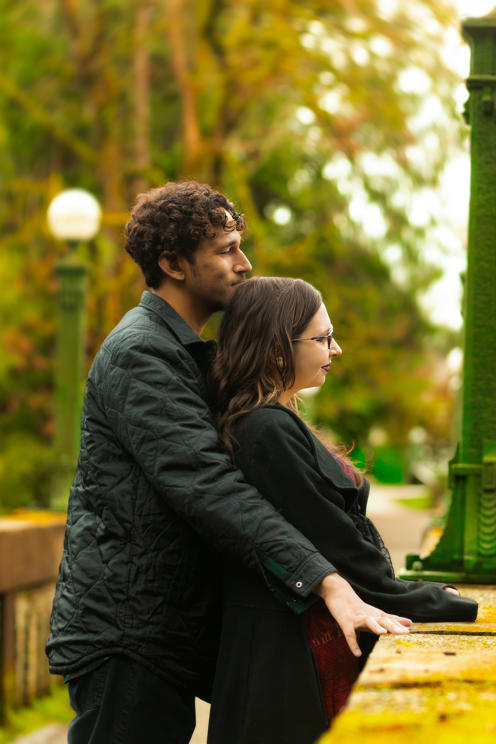 Two people lean against a railing in a park on an autumn day, wearing dark jackets while looking off into the distance.