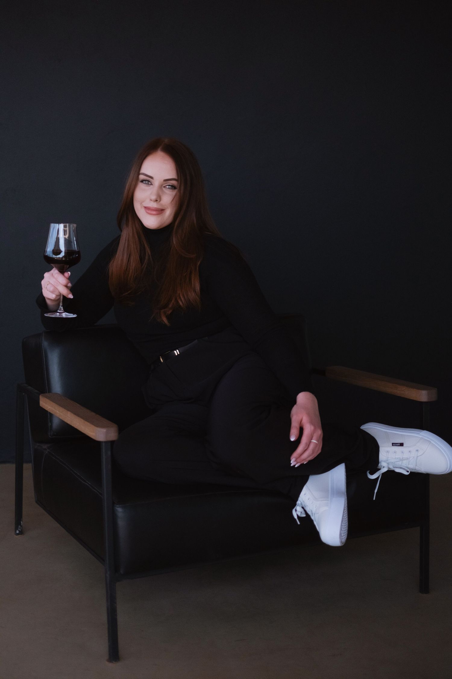 Woman in black outfit sits in leather chair holding glass of red wine.