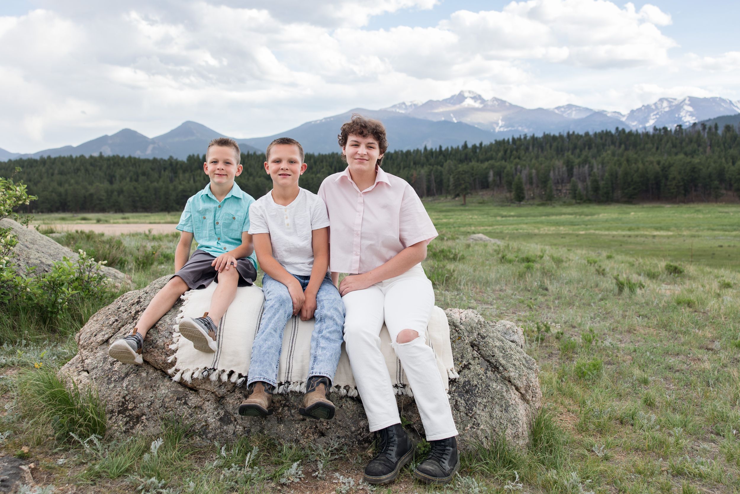 Three people sit together on a large rock with snow-capped mountains, forest, and meadow in the background.