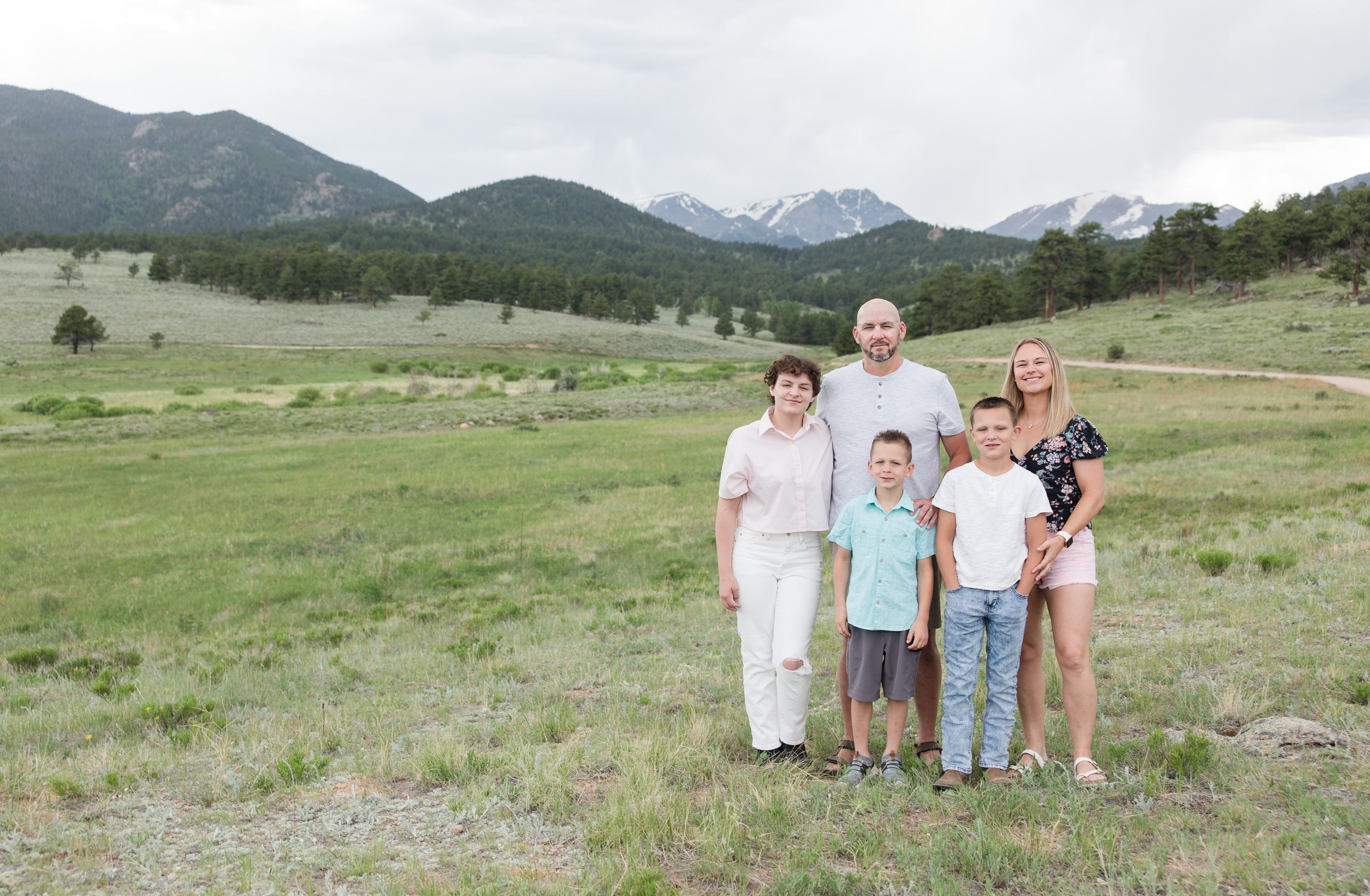 A family stands together in a scenic mountain meadow with snow-capped peaks and green hills in the background.