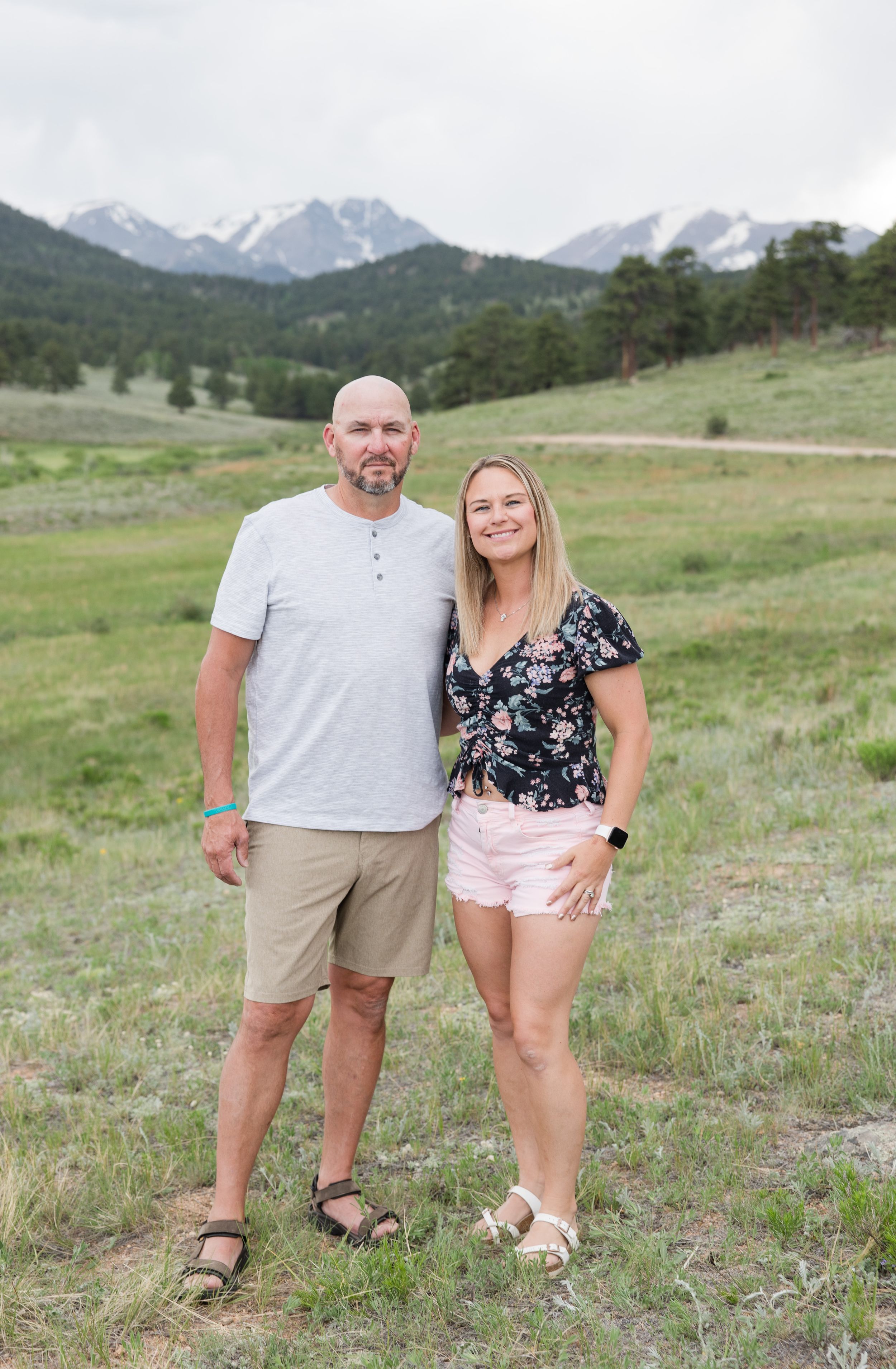 A couple stands together in a scenic mountain meadow with snow-capped peaks visible in the background on a sunny day.