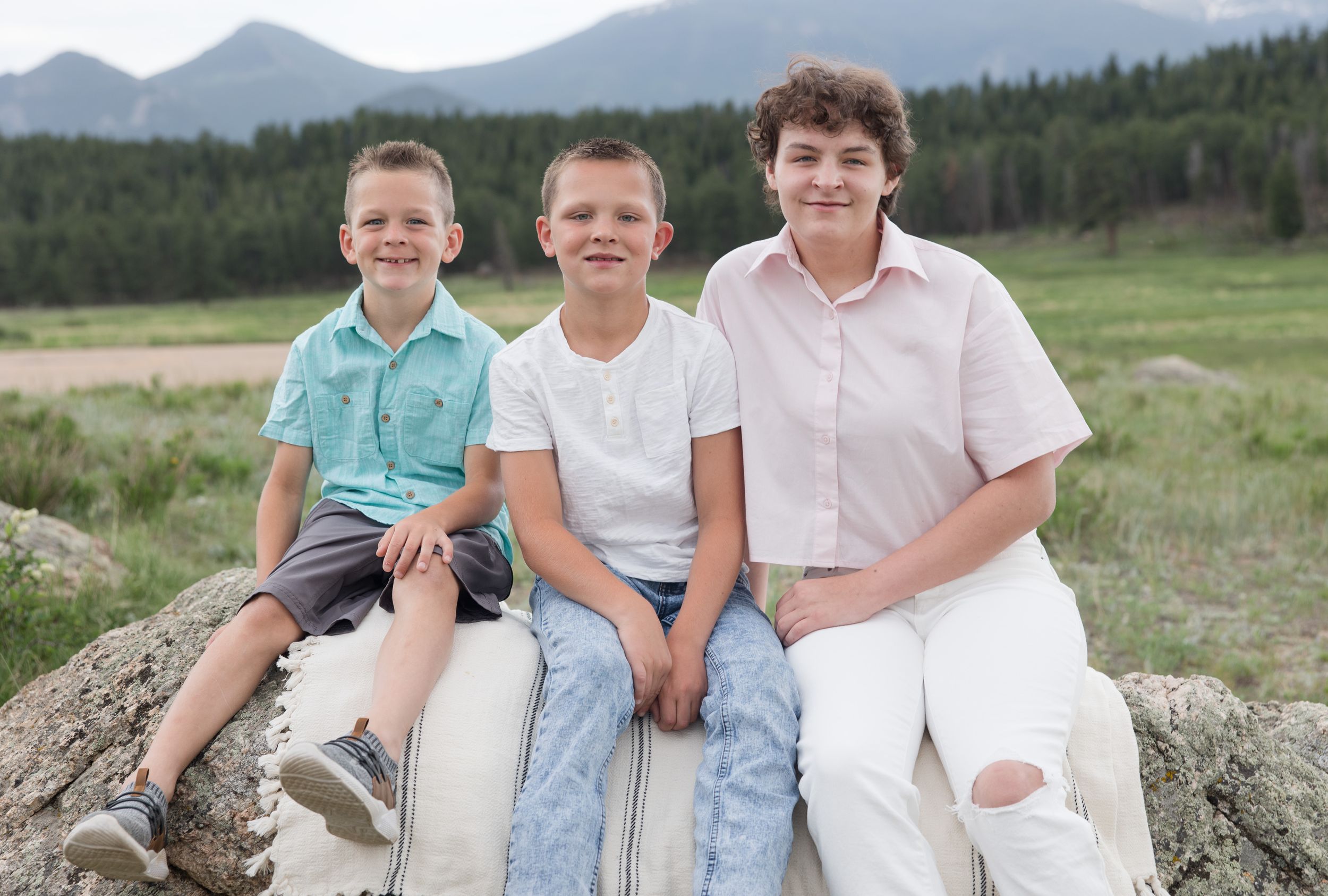 Three people sitting together on a large rock with mountain peaks and forest visible in the scenic background.