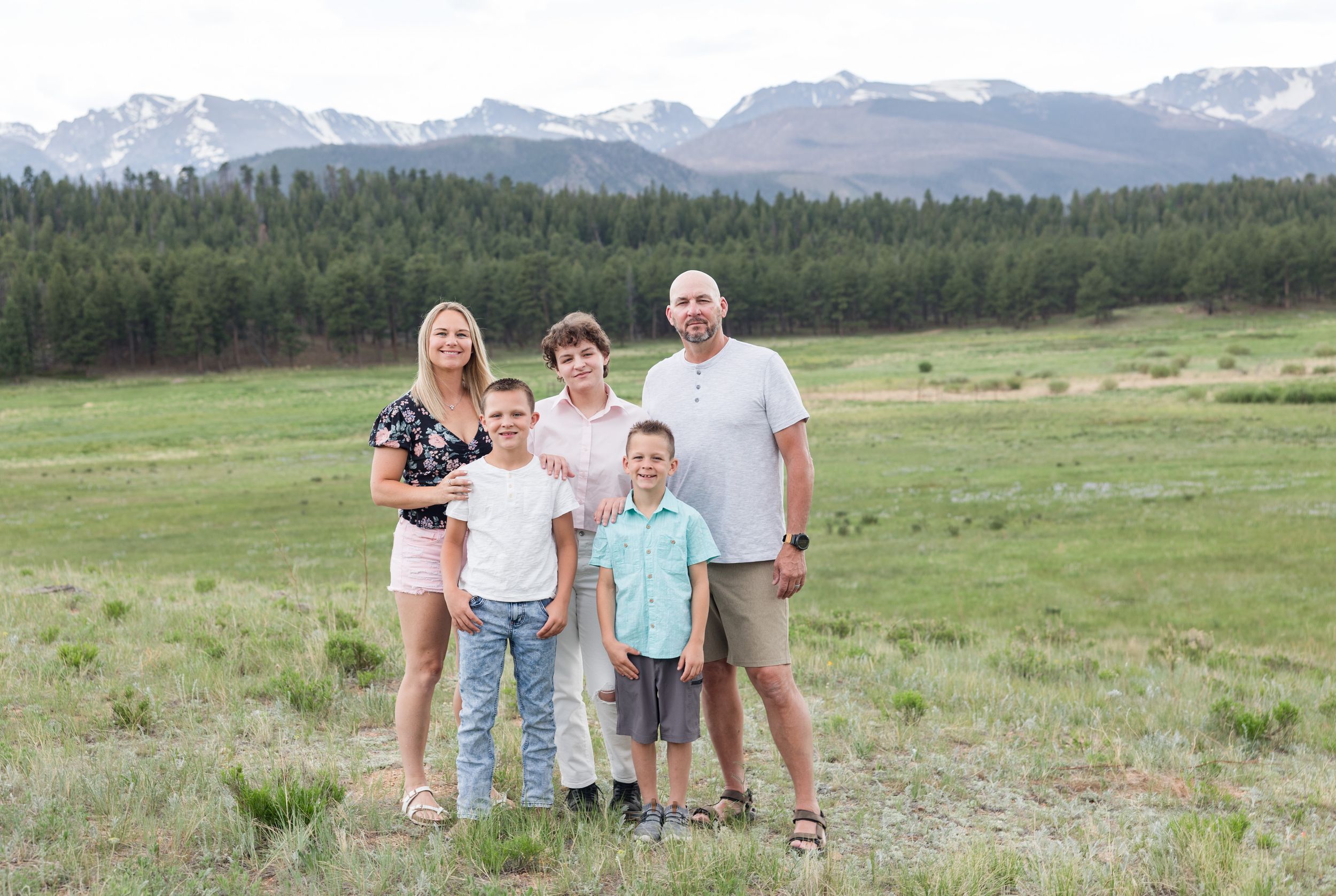 A family stands together in a green meadow with snow-capped mountain peaks and pine forest in the background.