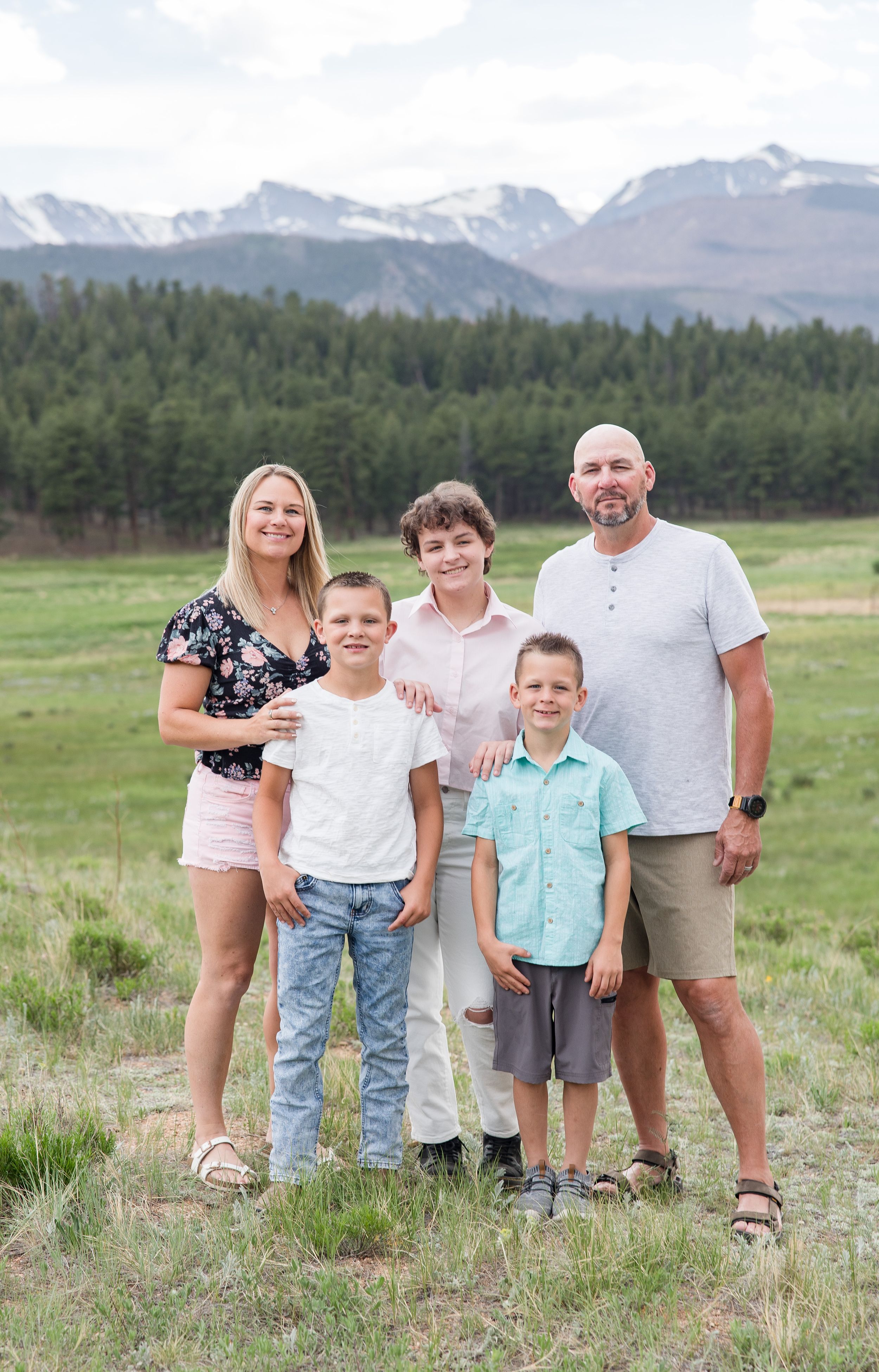 A group poses for a casual outdoor portrait with mountain peaks and evergreen forests in the scenic background.