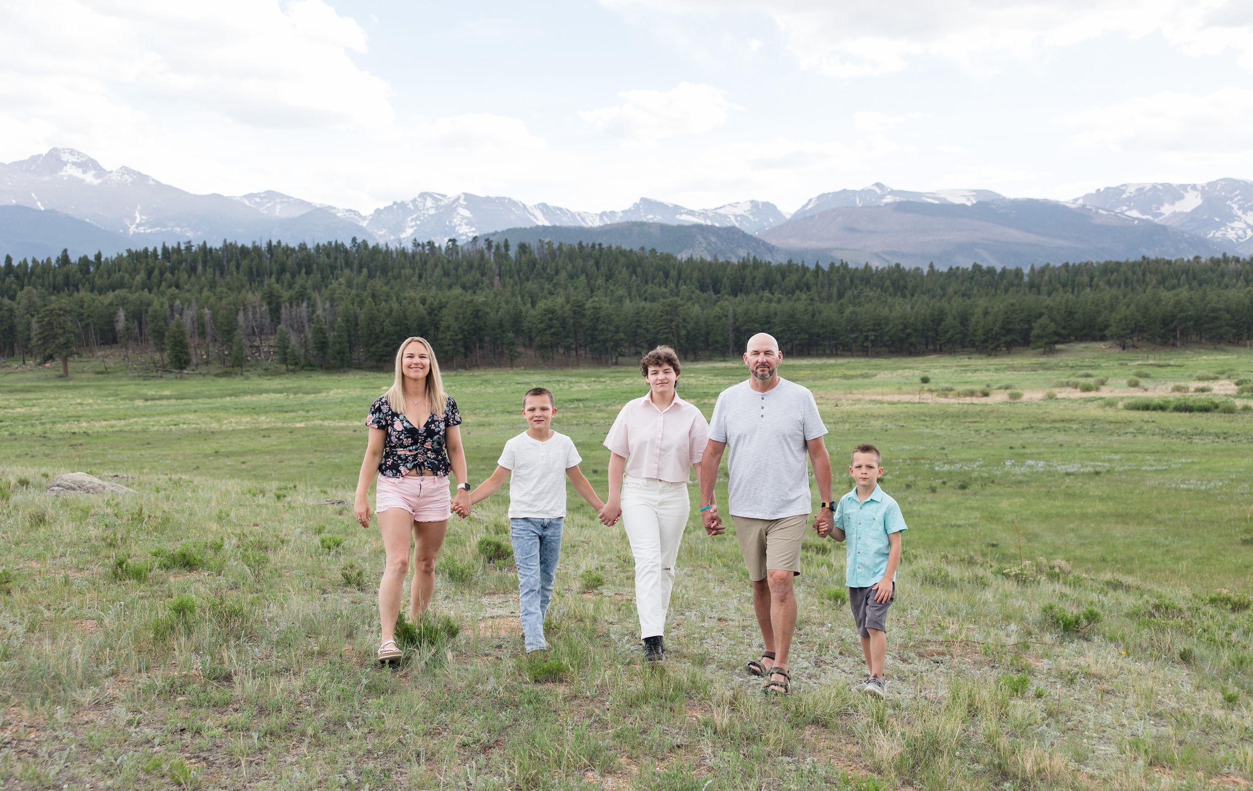 A group walks together across a green meadow with snow-capped mountains and a forest backdrop in the distance.