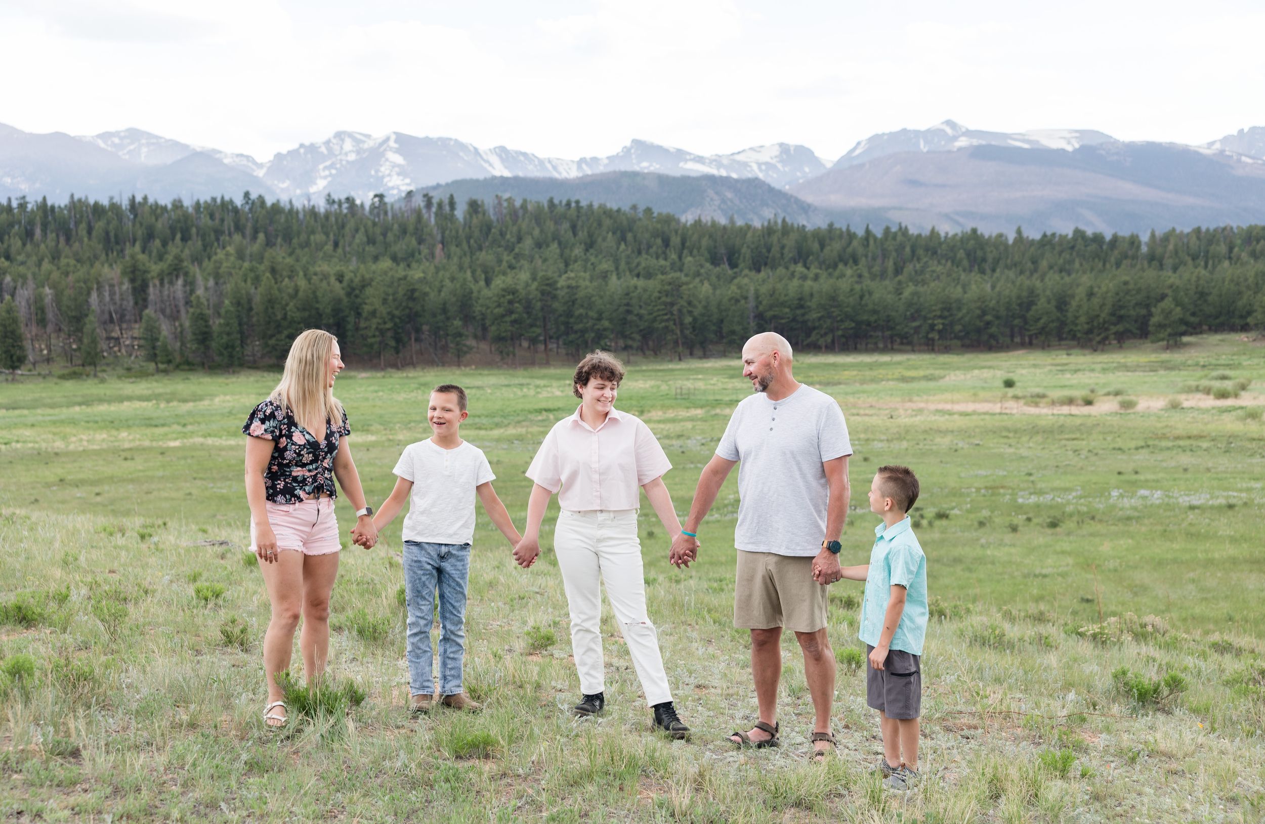 Family holds hands while standing in a mountain meadow with evergreen forest and snow-capped peaks in the background.