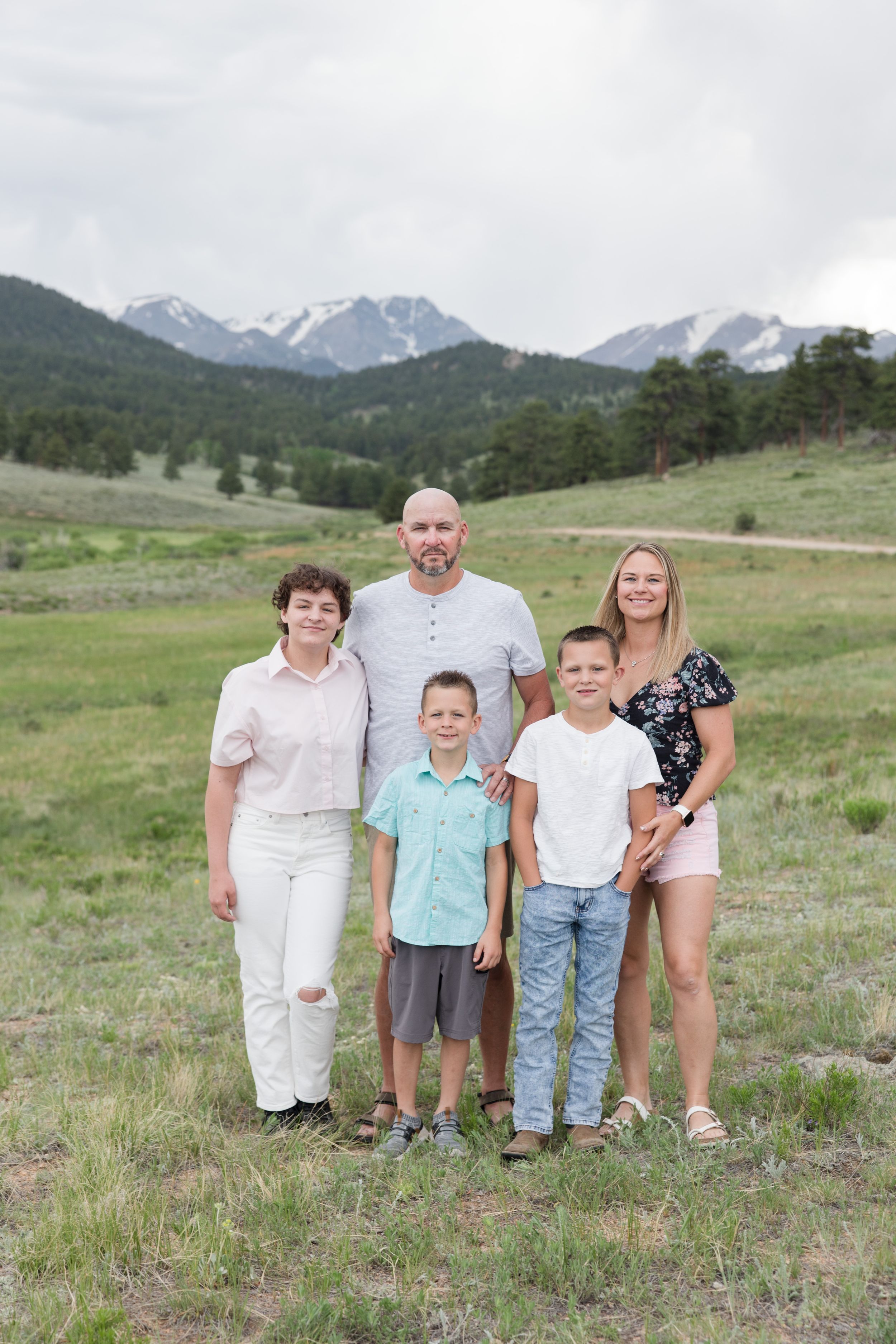 Family standing together in a grassy meadow with snow-capped Rocky Mountains visible in the distance.
