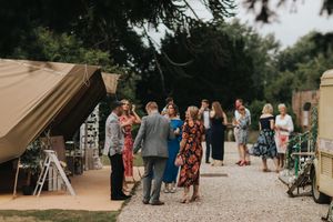 Wedding guests gather outside a tan tent for an outdoor reception on a gravel path surrounded by trees at Hirst Priory.