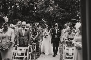 Black and white outdoor forest wedding ceremony with gathered guests seated along a leaf-strewn aisle path at Hirst Priory.
