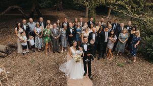 A wedding party of guests gathered in a rustic outdoor forest setting forming a group photo at Hirst Priory.