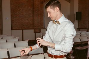 Individual in formal attire checking time on wristwatch while standing in auditorium with rows of chairs.