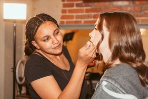 A makeup artist applies cosmetics on a client in a rustic brick-walled setting.