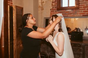 Wedding stylist adjusting bride's veil in rustic brick-walled room.