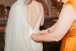 Orange-clad attendants helping with final adjustments to a wedding dress against brick wall backdrop.