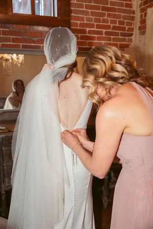 Someone helps button up the back of a white wedding dress with delicate veil detail in a brick-walled room.