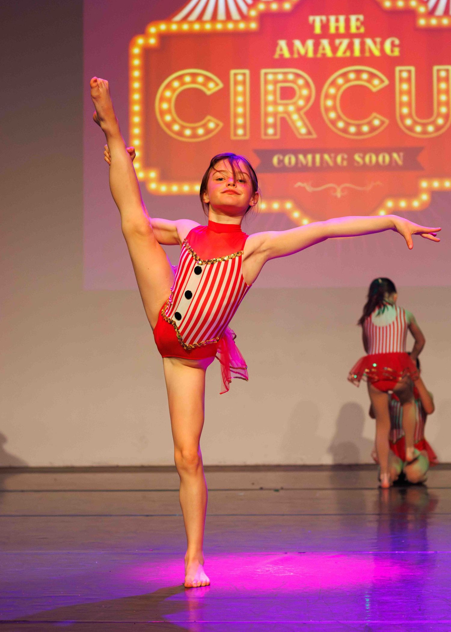 Young dancer performs upright split pose in red costume during circus-themed recital with colorful stage lighting.