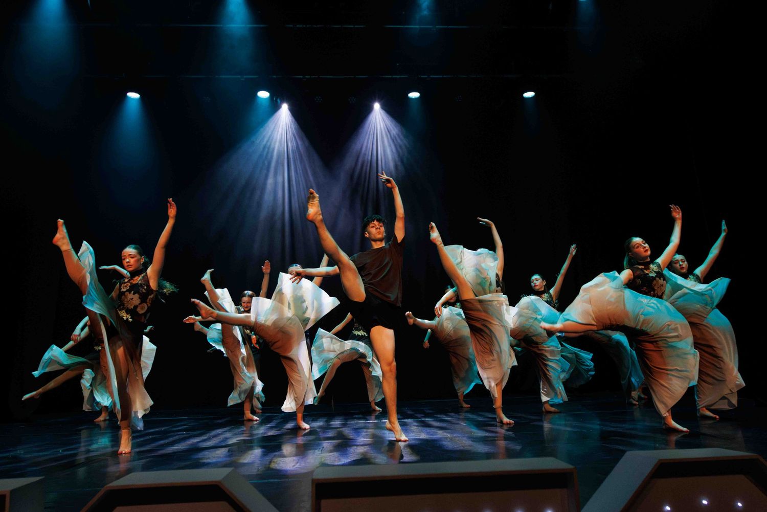 Dancers in turquoise and white costumes perform under dramatic blue spotlights on a dark theater stage.