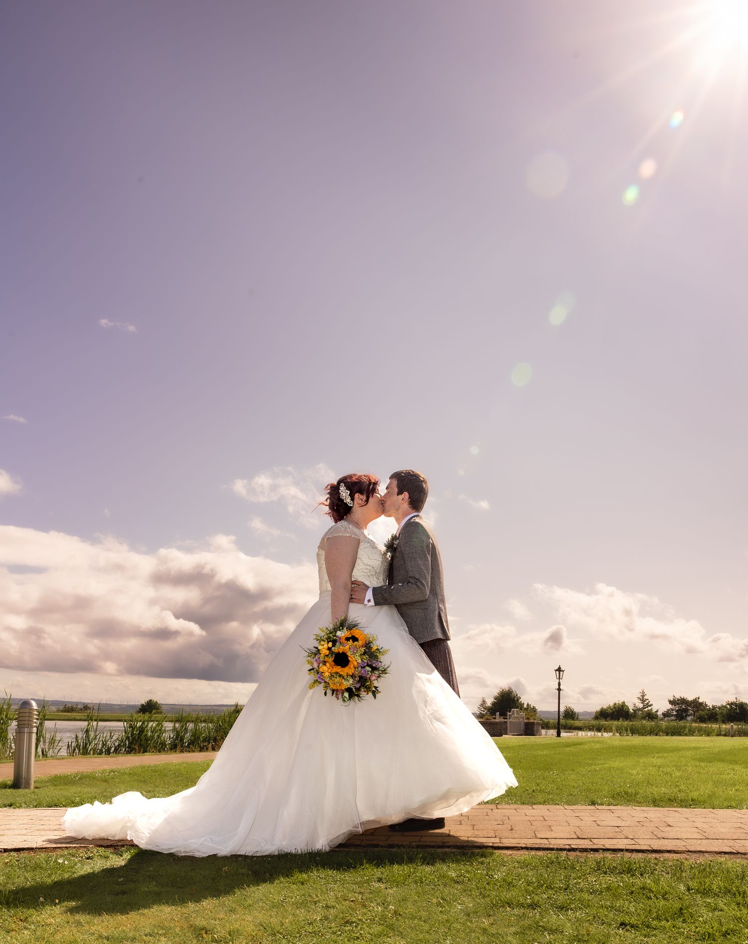 On a sunny day at an outdoor venue, a bride in a flowing white gown carries sunflowers with scenic landscaping in view.