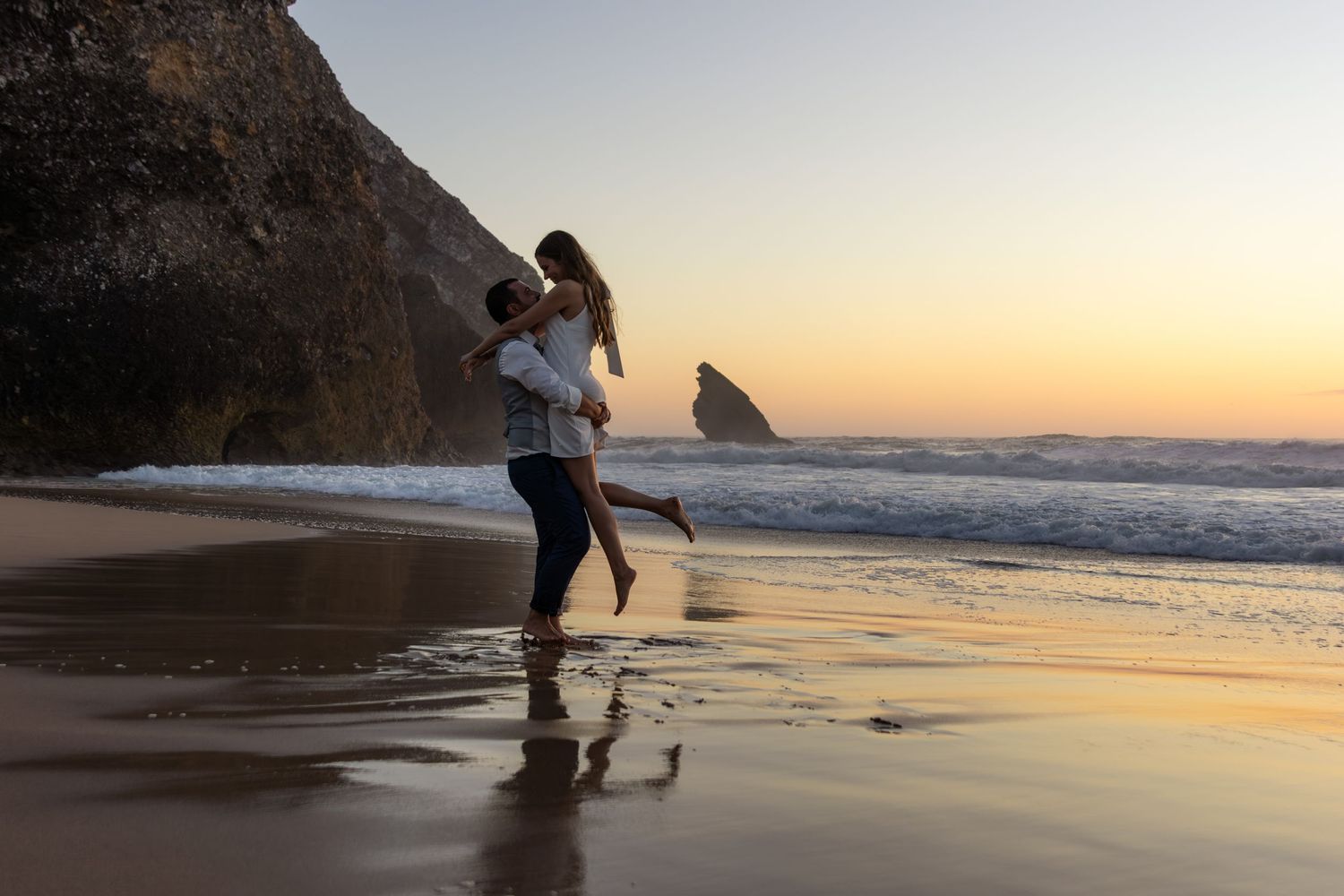 A couple shares intimate moments on a beach at sunset with waves crashing in the background.