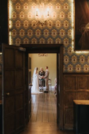 A wedding ceremony viewed through an open doorway with ornate wallpaper and wooden paneling visible in the foreground.