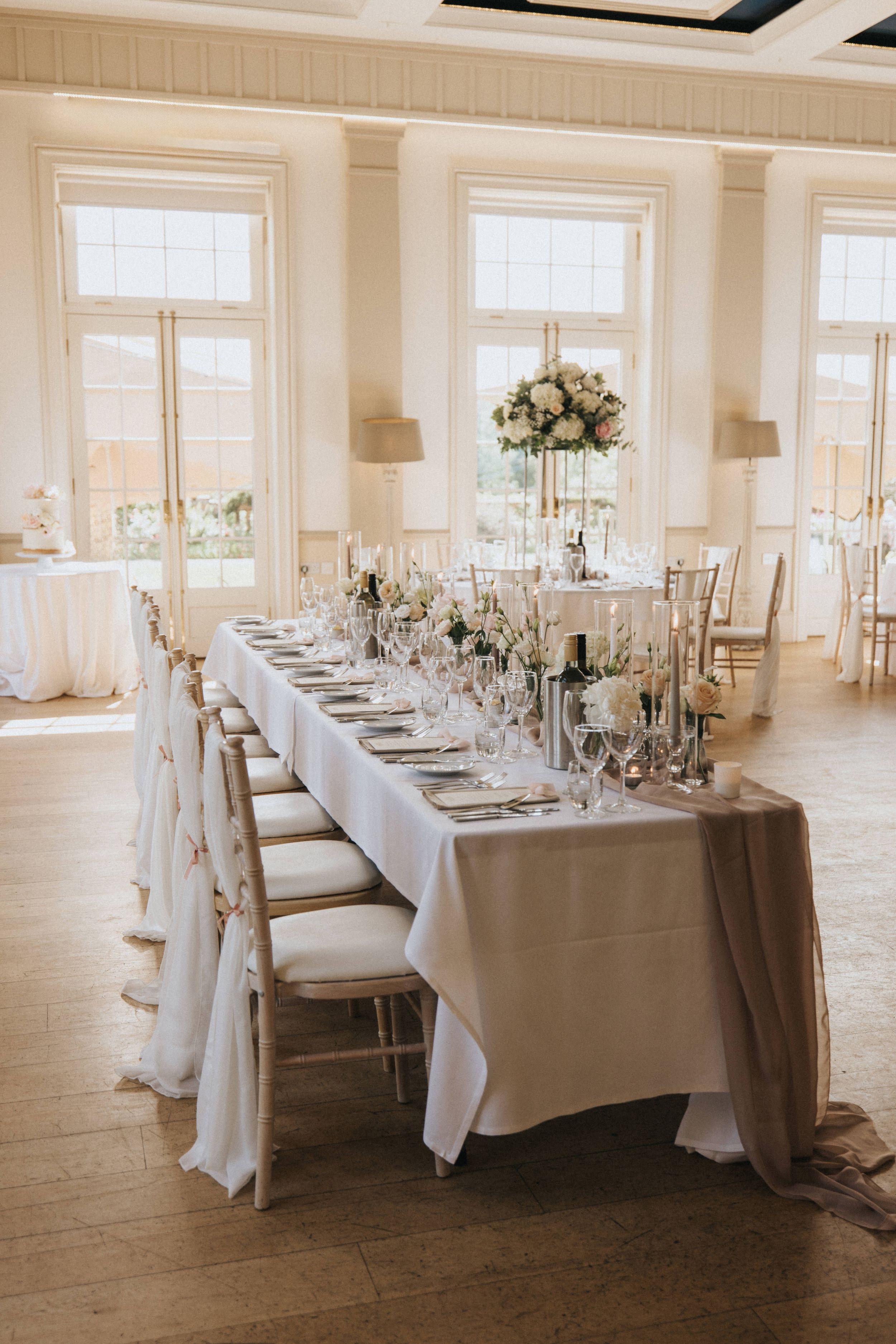 Elegant wedding reception table with white linens, floral centerpieces and crystal glassware set in a bright room with tall windows.