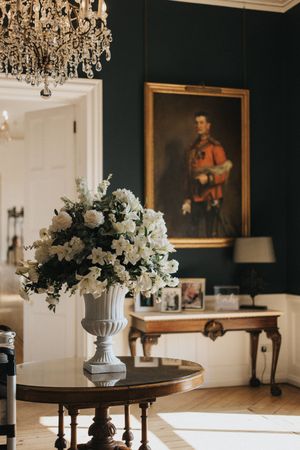 An elegant dining room with dark walls features crystal chandeliers, portrait paintings, and white floral centerpieces.