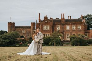 A couple shares a romantic wedding moment on the lawn of a majestic historic manor house with grand architecture.