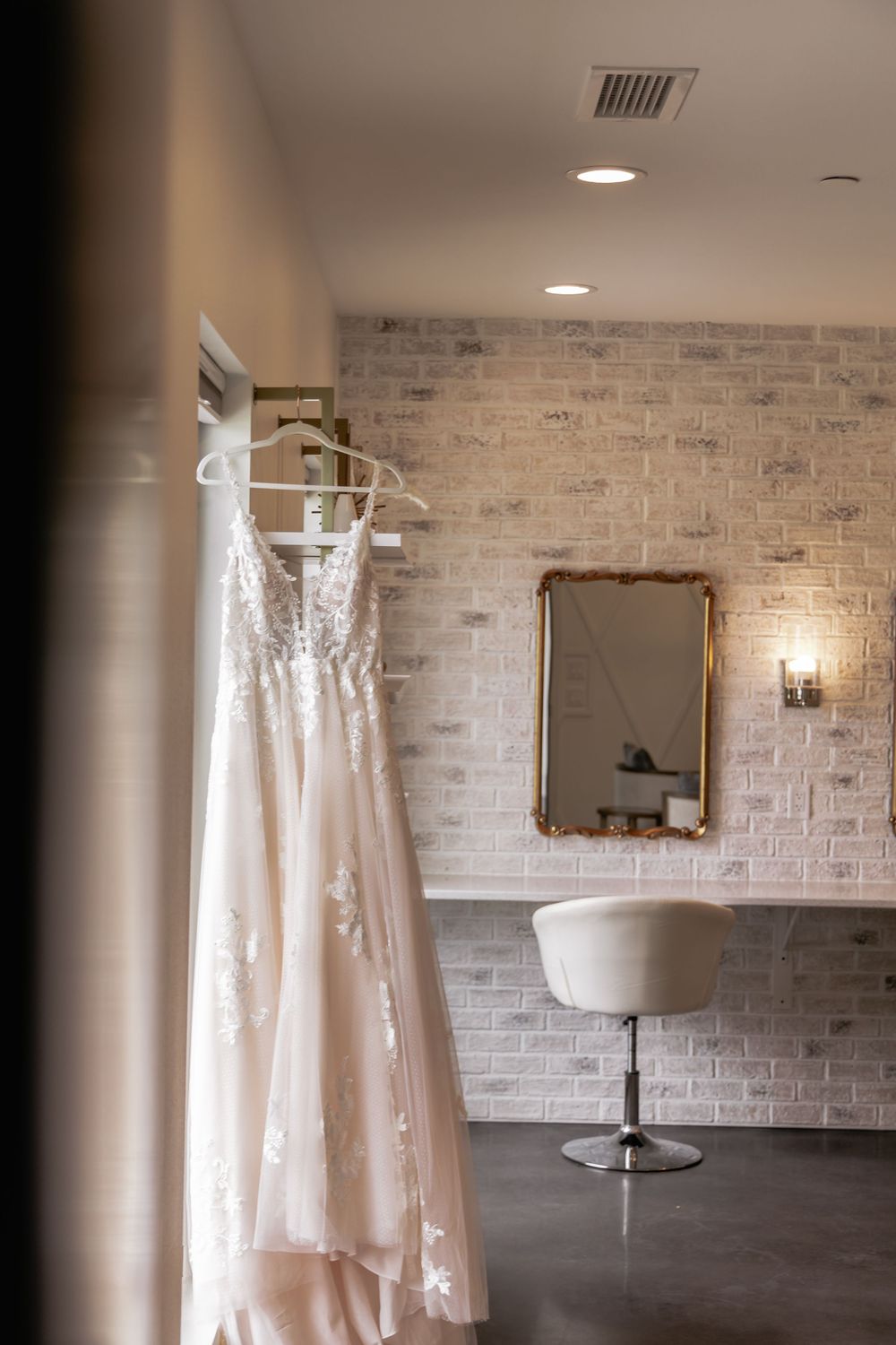 A blush wedding dress hangs in a bridal suite with exposed brick walls, a mirror, and modern decor.