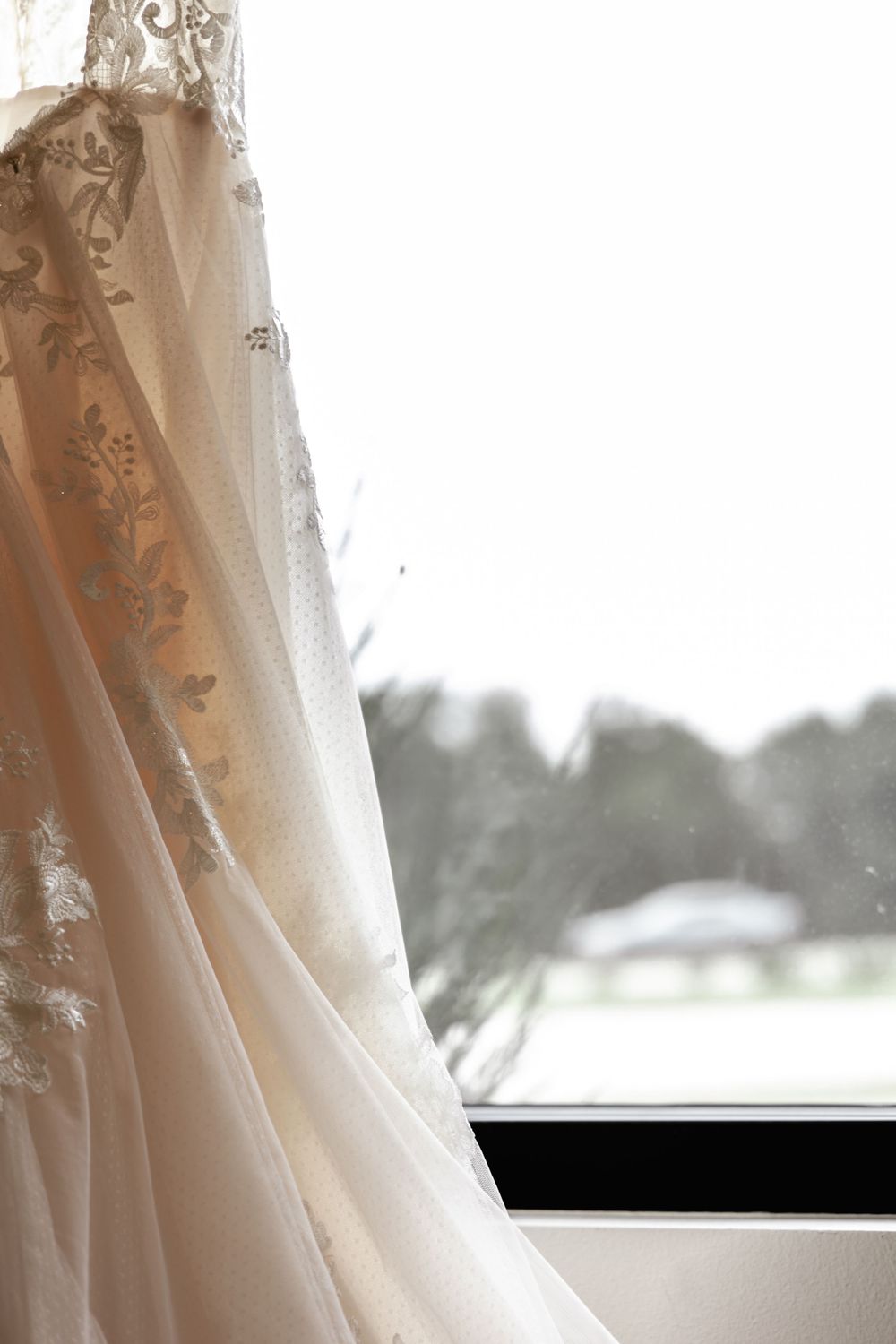Close-up detail of a white wedding dress with delicate lace trim hanging near a window in soft natural light.