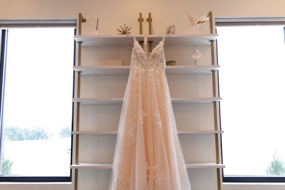 A delicate blush-colored wedding dress with lace details hangs elegantly between two windows on white shelving.