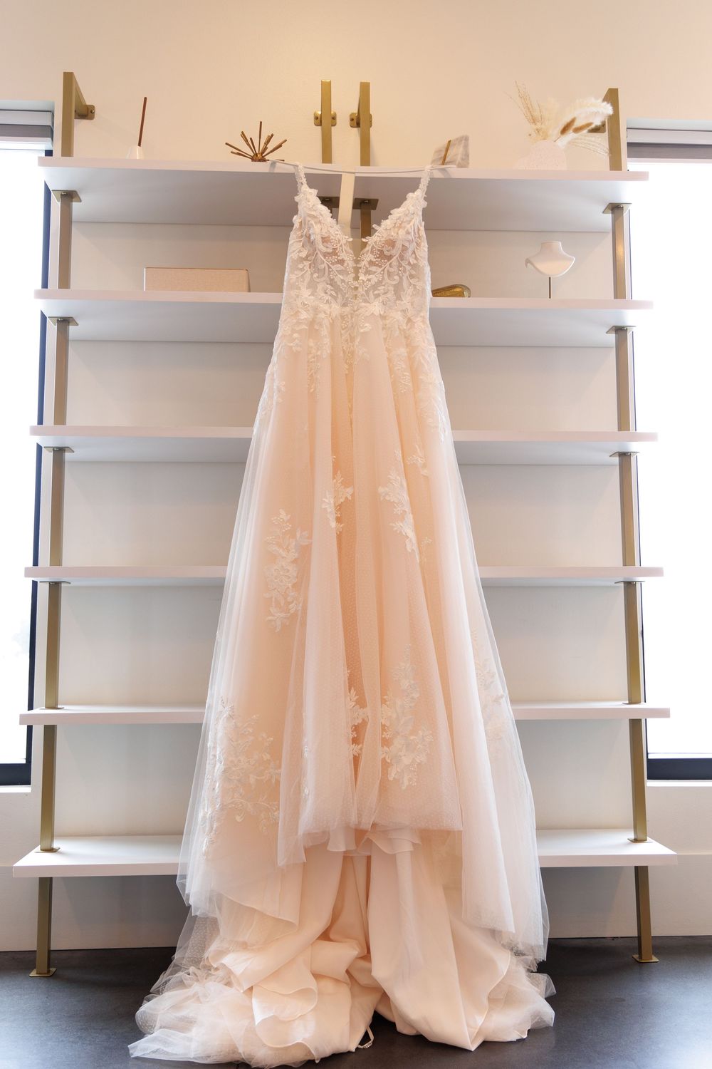 A blush colored lace wedding gown with delicate floral details hangs on display against white shelving.