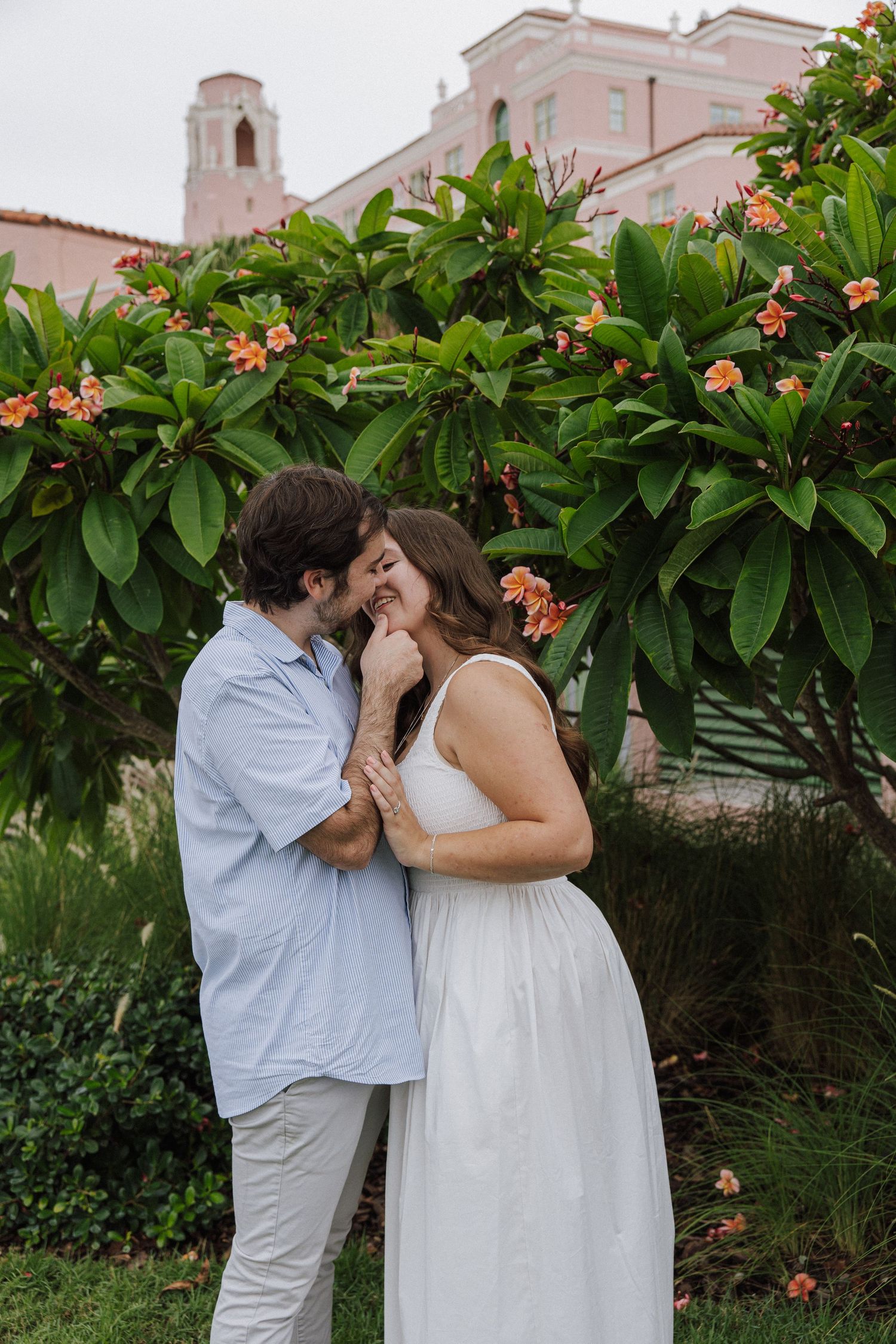 A couple shares an intimate moment beneath pink flowering bushes with a Spanish-style building in the background.