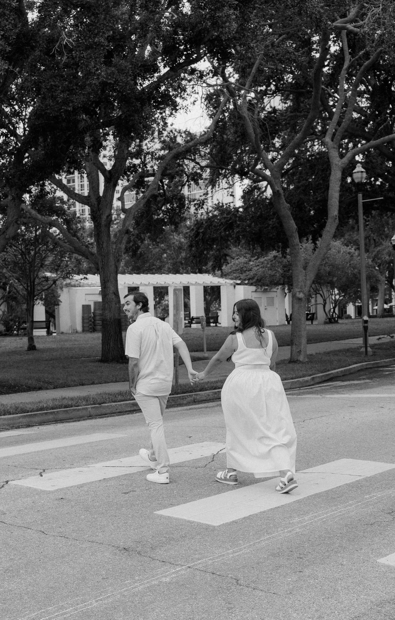 Four white, ghostly figures sit spaced apart in a line along a tree-lined street in a black and white photograph.