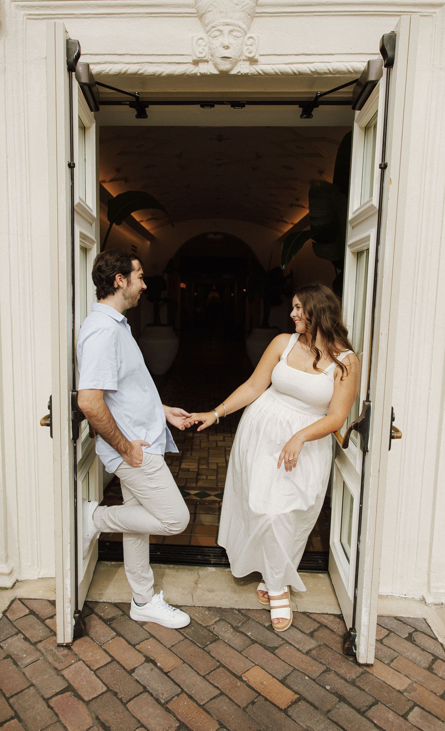 A couple holds hands in white outfits while standing in an open doorway of a brick-floored building.